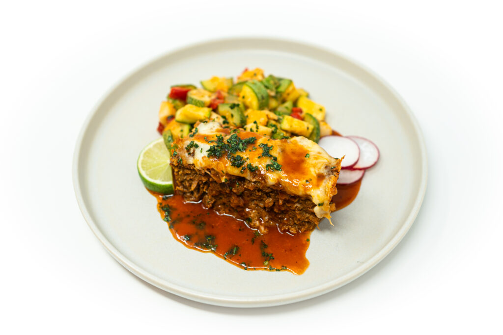 A slice of cheddar chorizo meatloaf drizzled with honey chipotle glaze, topped with melted cheese and crispy herbs, served with a fresh zucchini salad, radish slices, and a lime wedge on a neutral plate.