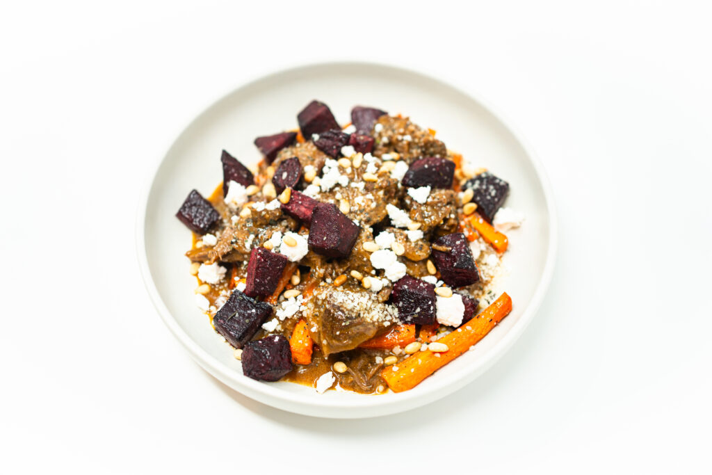 A plated dish of honey mustard braised lamb with roasted beets and carrots, garnished with crumbled goat cheese, toasted pine nuts, and a drizzle of reduced braising sauce, served on a minimalist white plate against a clean, bright background.