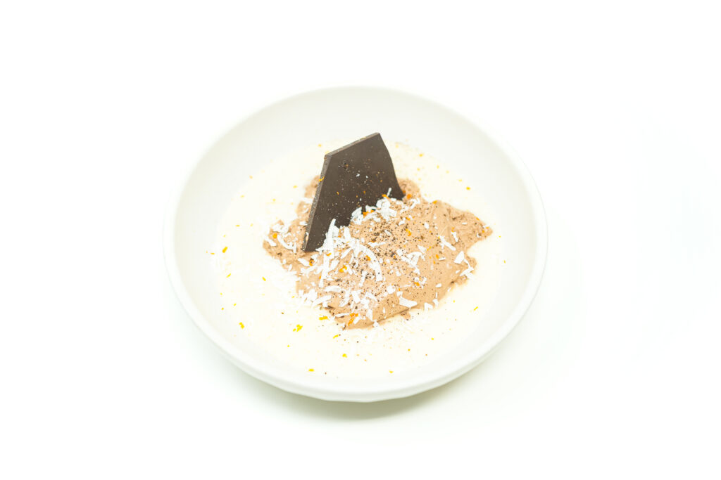 A dark chocolate mousse quenelle set over whipped coconut cream, topped with chili powder, toasted coconut flakes, and chocolate bark.