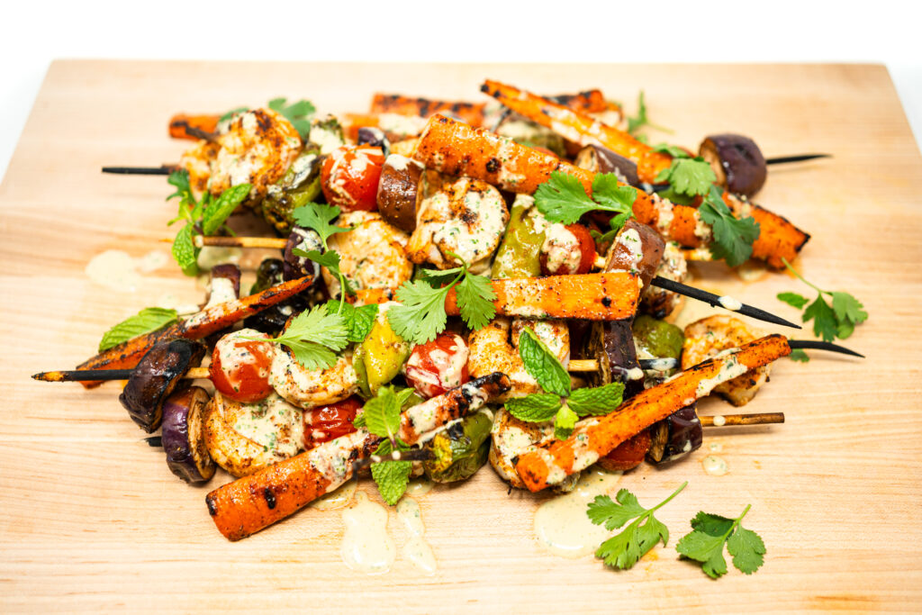 Grilled shrimp skewers stacked on a wooden board with blistered shishito peppers, cherry tomatoes, Indian eggplant, and roasted carrots, all drizzled with herbed coconut cream and garnished with fresh cilantro and mint.