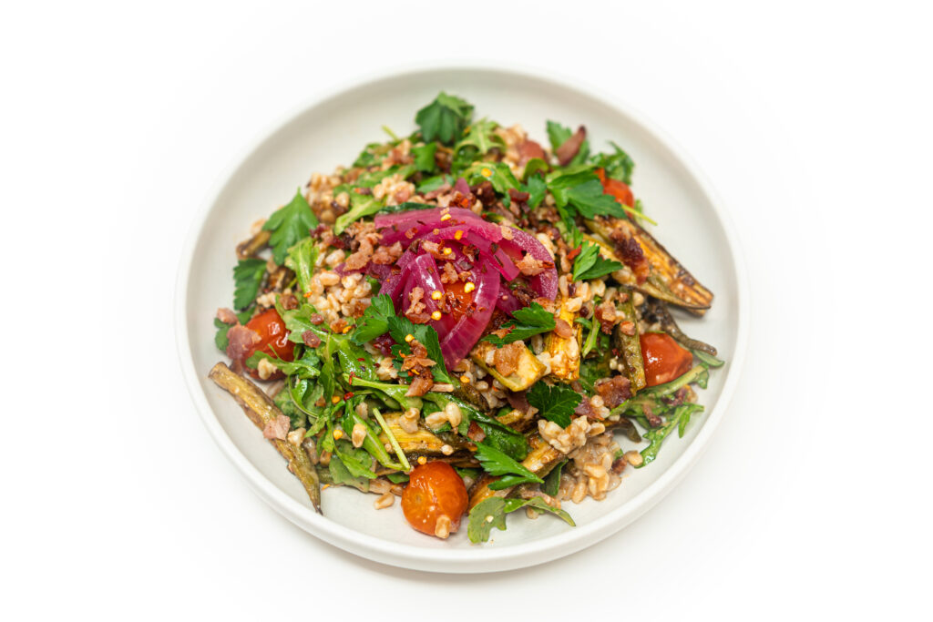 A vibrant bowl of okra farro salad topped with arugula, cherry tomatoes, parsley, pickled red onions, and bacon vinaigrette.