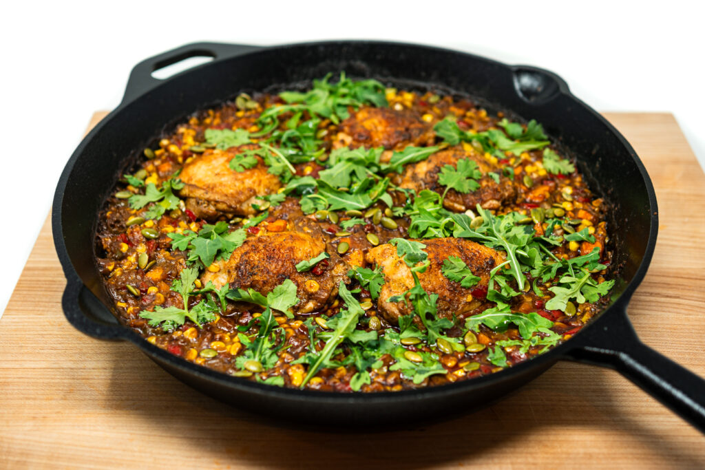 Crispy chicken thighs simmered in a mole-infused broth with corn and serrano peppers, topped with arugula and toasted pepitas in a cast iron skillet.