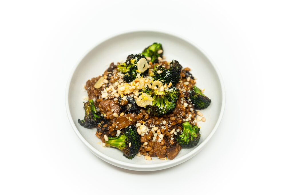 Overhead view of a white bowl filled with pearl couscous tossed with wild mushrooms and sun-dried tomatoes, topped with roasted broccoli, toasted pine nuts, and grated Parmesan cheese on a clean white background.