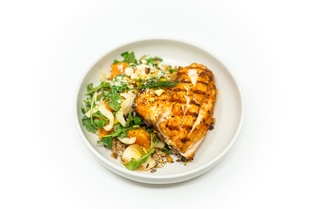 Grilled striped bass fillet served with charred citrus, grilled fennel, endive, and arugula salad on a white plate.