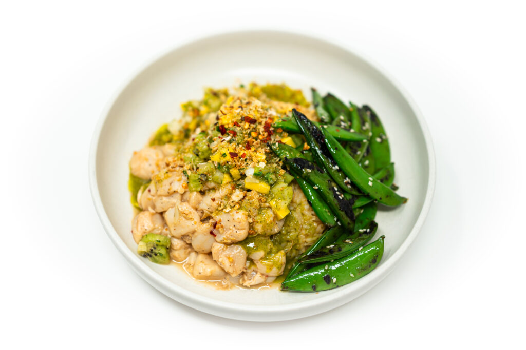 Scallop and escolar ceviche served in a white bowl, topped with kiwi-mango salsa and garnished with herbs and crushed spices, alongside charred sugar snap peas.