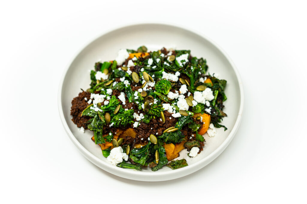 A bowl of miso ground beef with roasted butternut squash, charred rapini, crumbled goat cheese, toasted pepitas, and a drizzle of honey.
