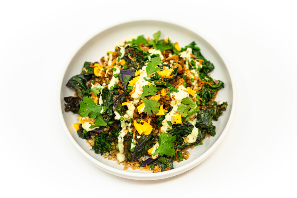 Lemongrass ground chicken bowl with cauliflower rice, charred kale, diced mango, and a spoonful of avocado lime crema, served in a shallow white bowl.