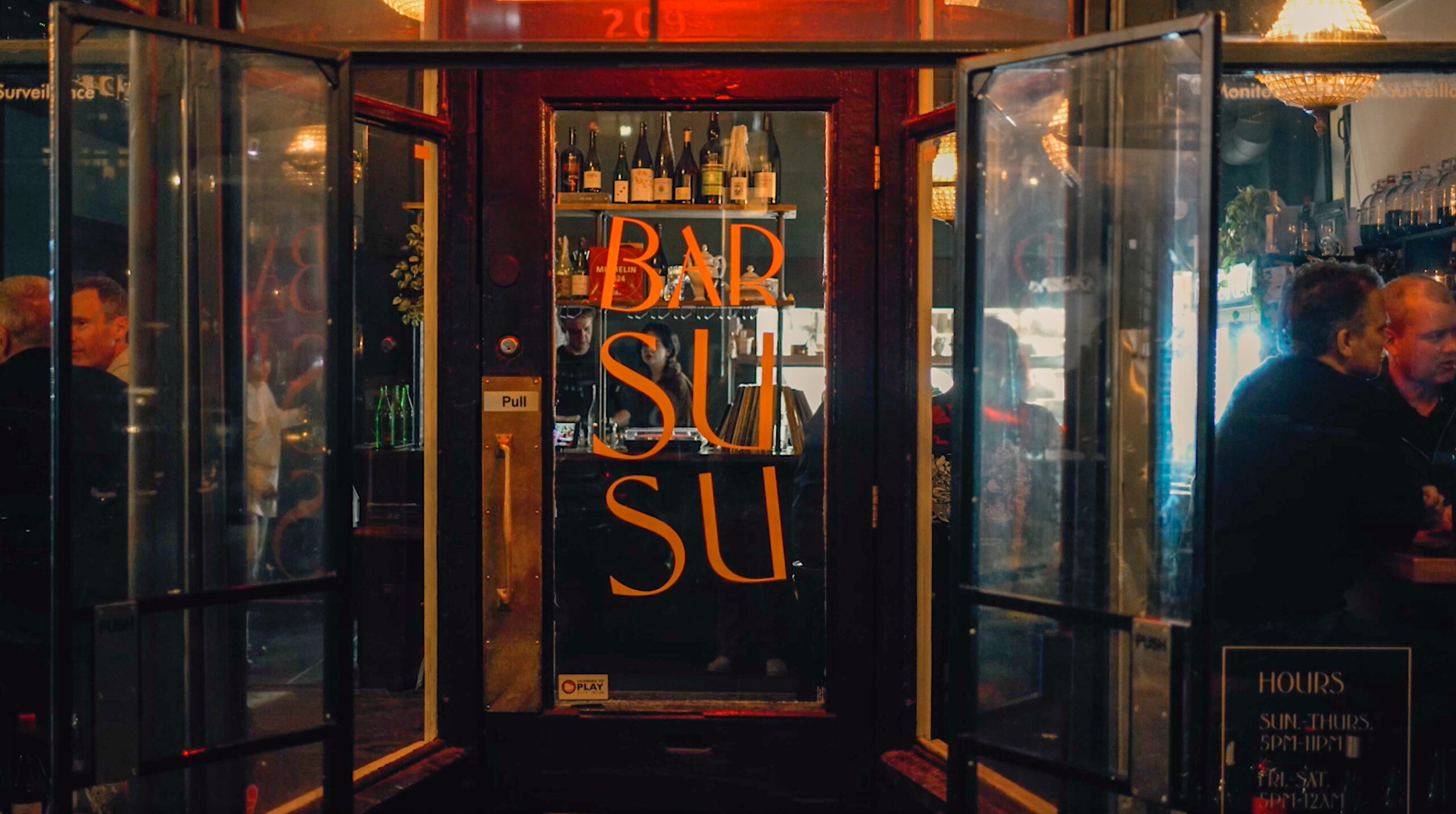 Entrance to Bar Susu in Vancouver, with glowing gold signage and patrons visible through the glass doors.