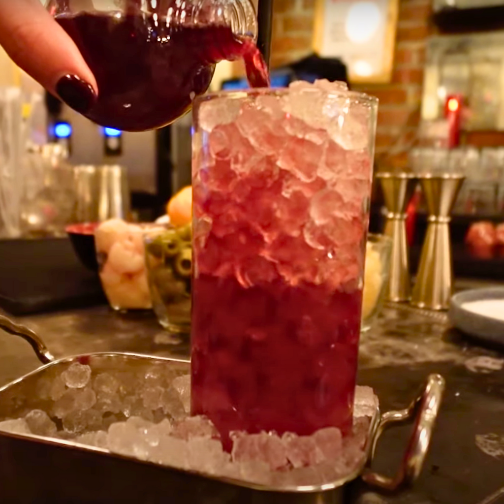 Tall glass being filled with a purple cocktail over crushed ice, surrounded by tropical drink ingredients.