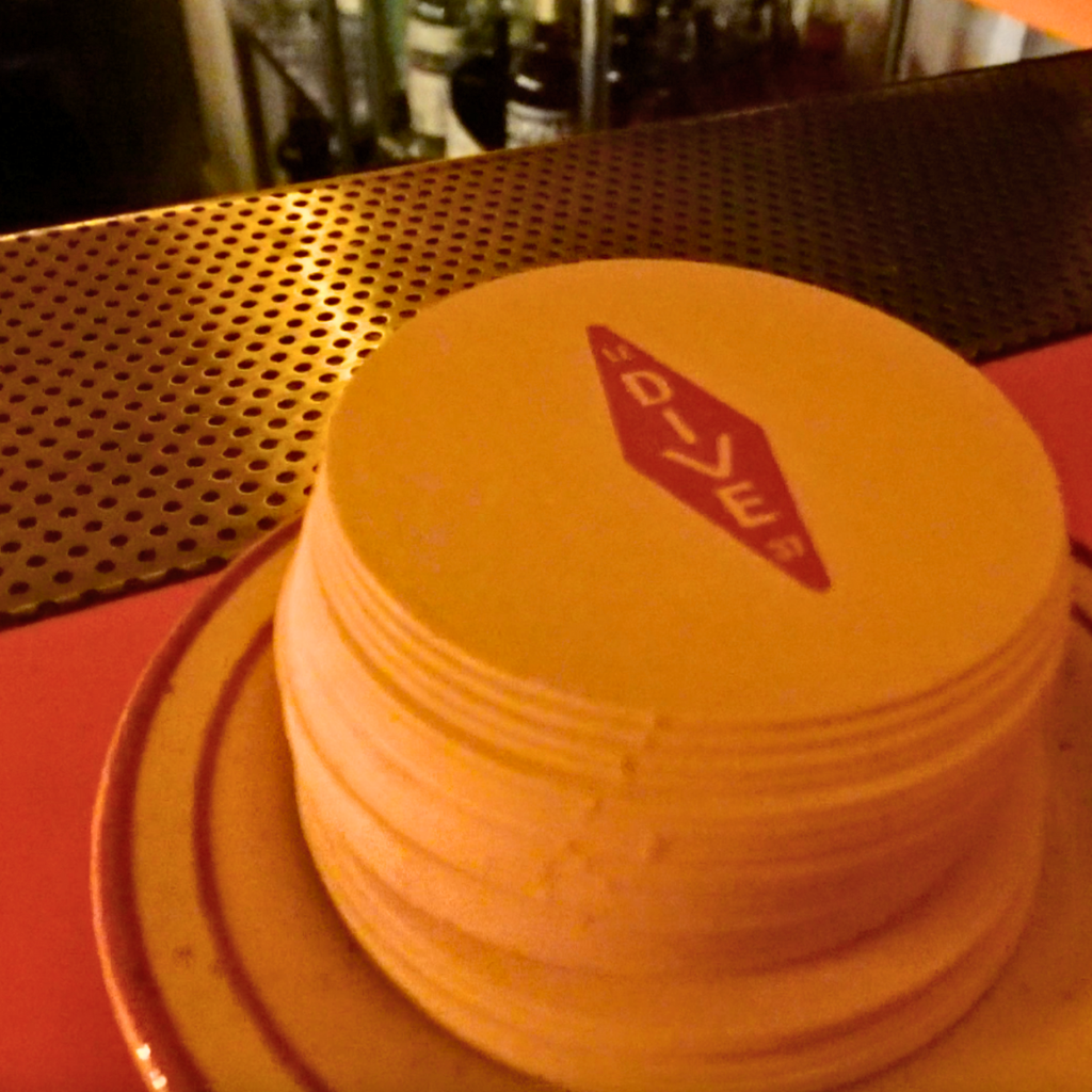 Stack of Le Dive coasters on a bar counter, with warm lighting and a perforated brass surface behind.