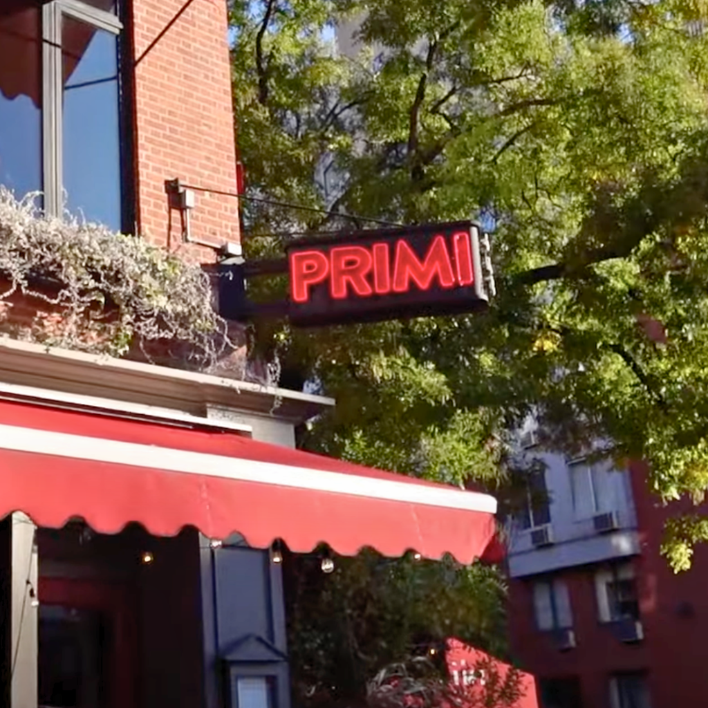 Exterior of Bar Primi with red awning, neon sign, and leafy trees along the Bowery.