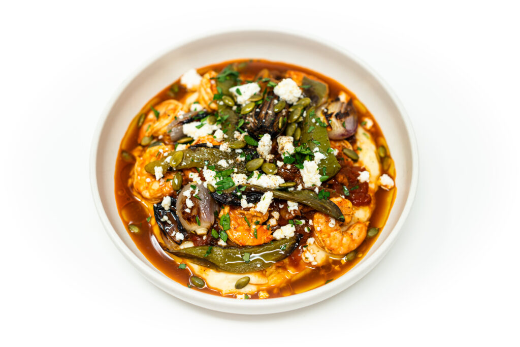 Bowl of creamy masa polenta topped with spiced shrimp, charred peppers and shallots, garnished with cotija cheese, toasted pepitas, cilantro, and lime wedges.