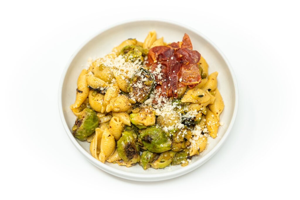 A white bowl filled with Brussels Sprouts Carbonara made with pasta shells, roasted Brussels sprouts, crispy prosciutto, grated Parmesan, and a creamy miso-butter sauce.