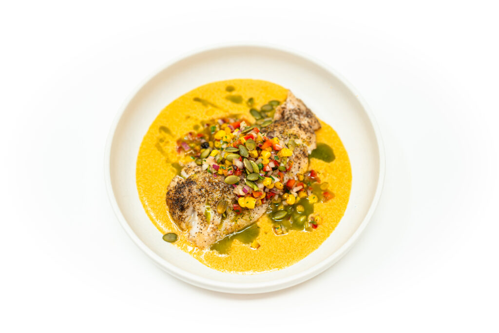Seared tilefish fillet topped with vibrant corn salsa, served over a creamy coconut corn sauce and finished with cilantro oil, plated in a shallow white bowl.