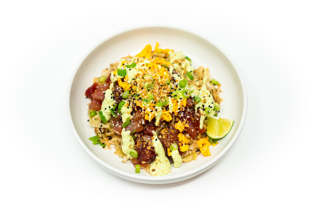 A vibrant tuna poke bowl with charred cabbage, diced mango, avocado crema, scallions, sesame seeds, and lime wedges on a white plate.