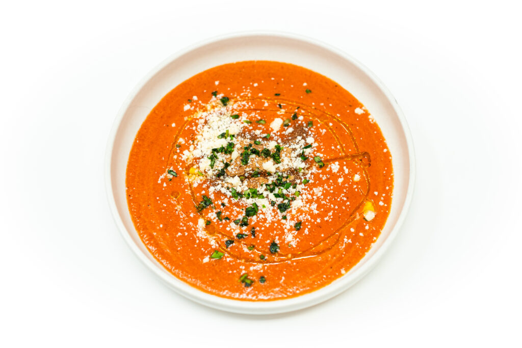 A chilled bowl of purple tomato and roasted red pepper gazpacho, topped with golden toasted breadcrumbs, fresh basil, and a drizzle of olive oil.