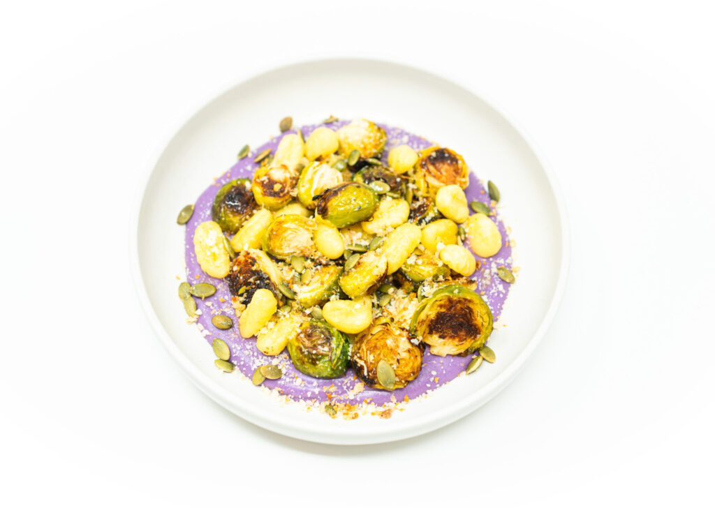 Crispy gnocchi served over a smooth purple ube purée, with seared Brussels sprouts, toasted pepitas, and lemon zest garnish on a ceramic plate.