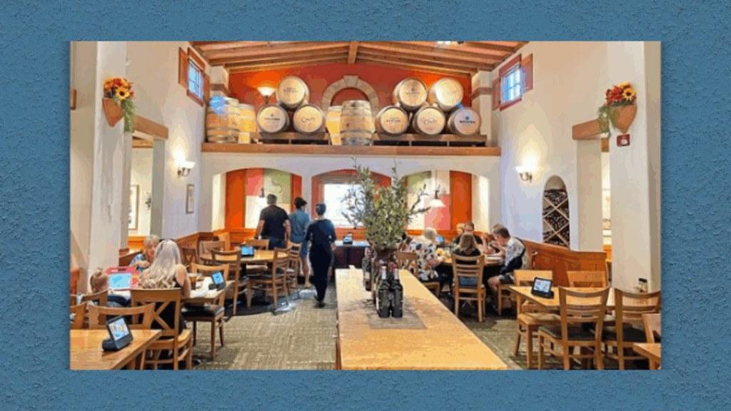 Interior of an Olive Garden restaurant with wooden tables, wall-mounted wine barrels, and diners seated in a casual Italian-style setting.