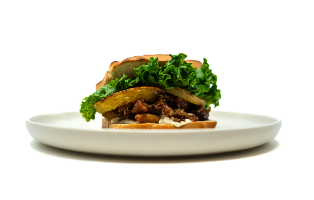 Close-up of a pork belly BLT with roasted green tomatoes, crisp lettuce, and lemon aioli on toasted bread.