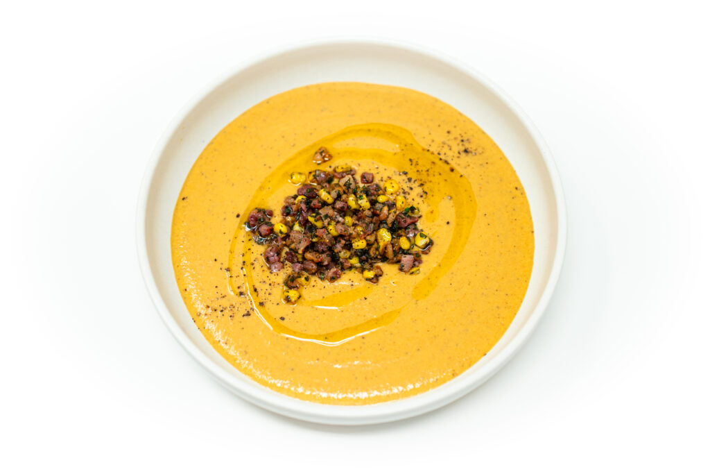 Chilled corn bisque topped with crispy pancetta and fresh parsley, served in a white bowl with a smooth, creamy texture.