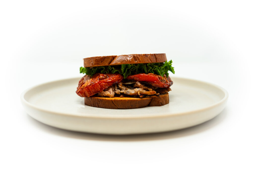 Close-up of a pork belly BLT with balsamic-marinated roasted tomatoes, crisp lettuce, and toasted bread on a white plate.