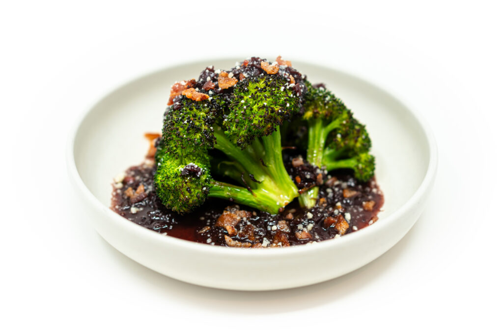 Charred broccoli crowns topped with red wine shallot bacon sauce and Parmesan on a white plate