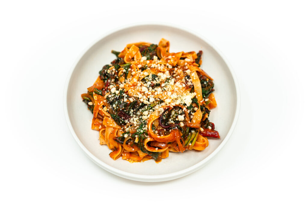 A bowl of tagliatelle pasta tossed with roasted rapini, wilted red chard, and a rich tomato sauce, topped with Parmesan and black pepper.