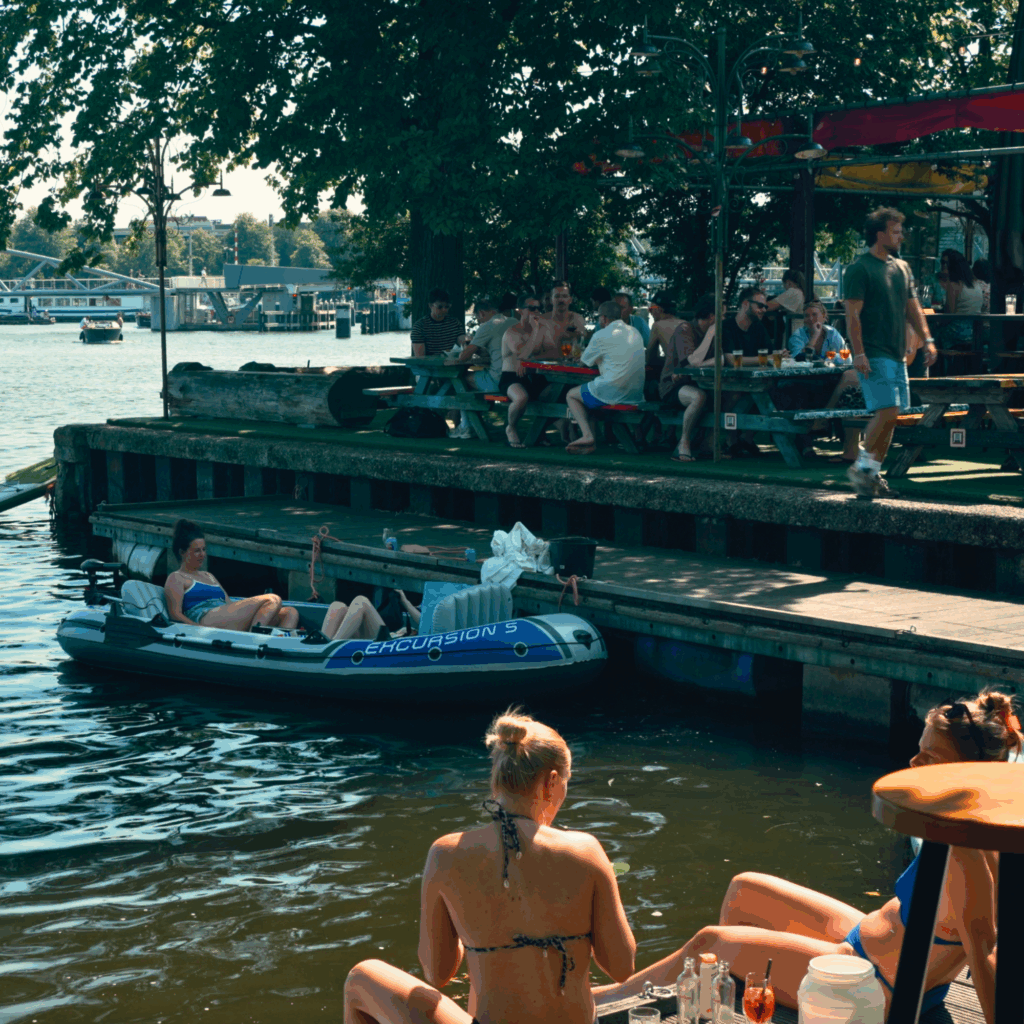 Hannekes Boom in Amsterdam, a lively waterfront bar with people sitting along the dock in the summer sun.