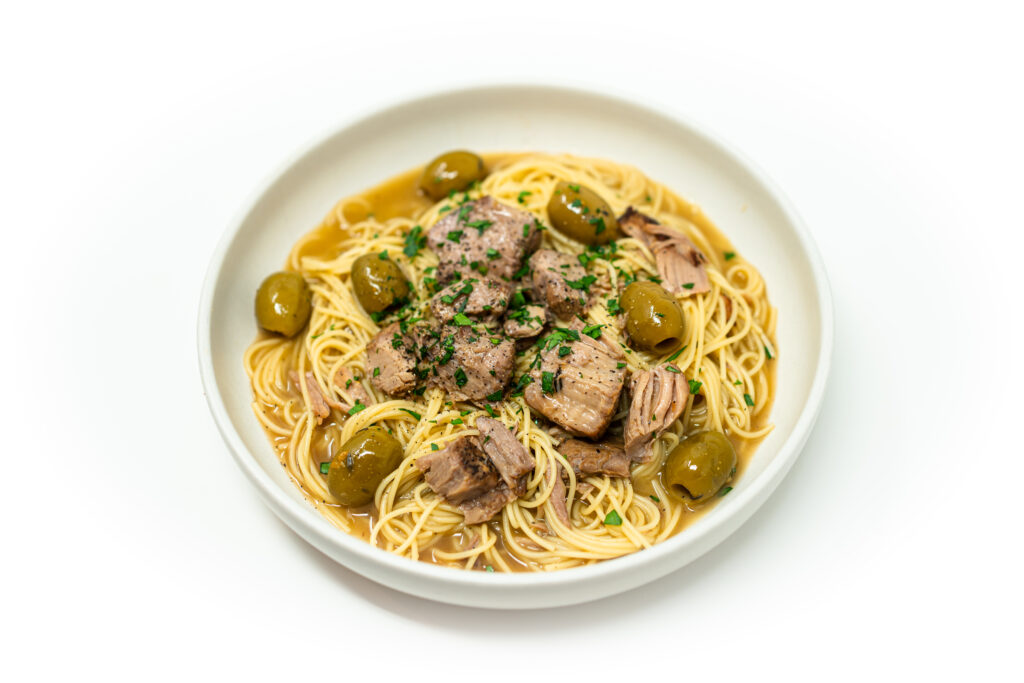 A bowl of capellini pasta topped with citrus braised veal shank, green olives, and fresh parsley in a golden broth.