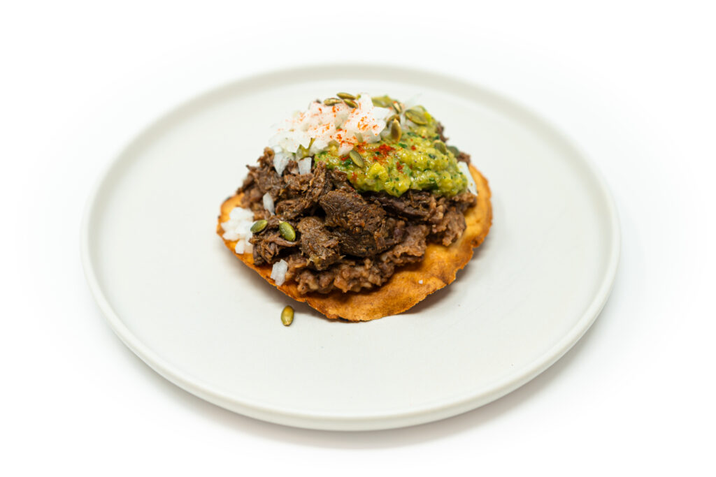 Crisp tostada topped with braised lamb, refried beans, charred jalapeño salsa, toasted pepitas, and diced onion on a rustic plate.
