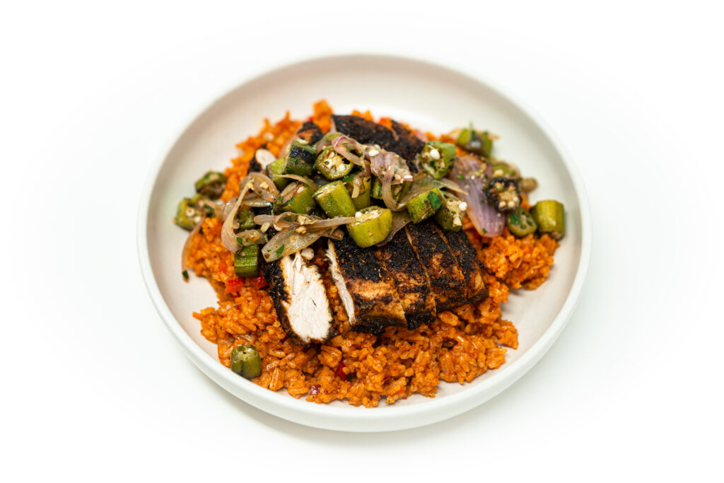 A plate of sliced blackened chicken served over seasoned tomato rice with charred okra and shallots on the side.