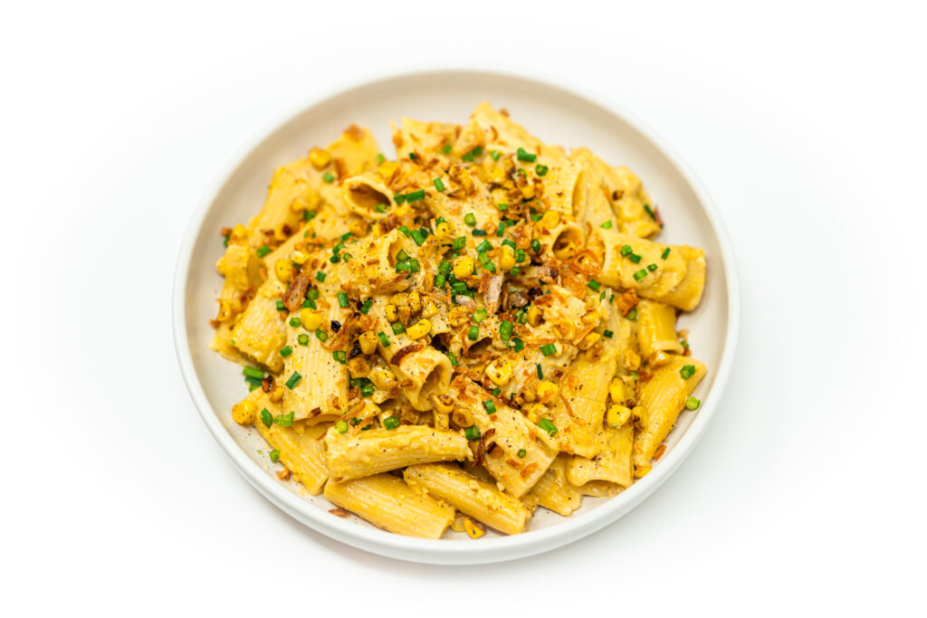 A bowl of rigatoni pasta coated in creamy corn carbonara sauce, topped with charred corn kernels, fresh chives, and grated Parmesan.