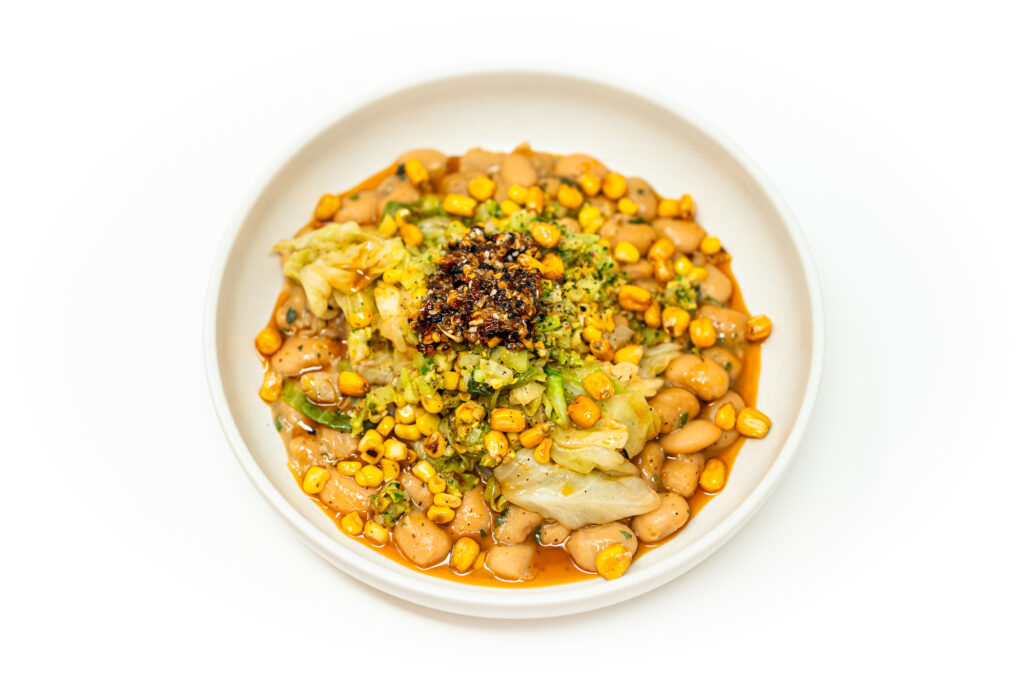 A bowl of slow-cooked cannellini beans topped with wok-charred cabbage and corn, finished with homemade chili crisp for a spicy, smoky vegetarian dinner.