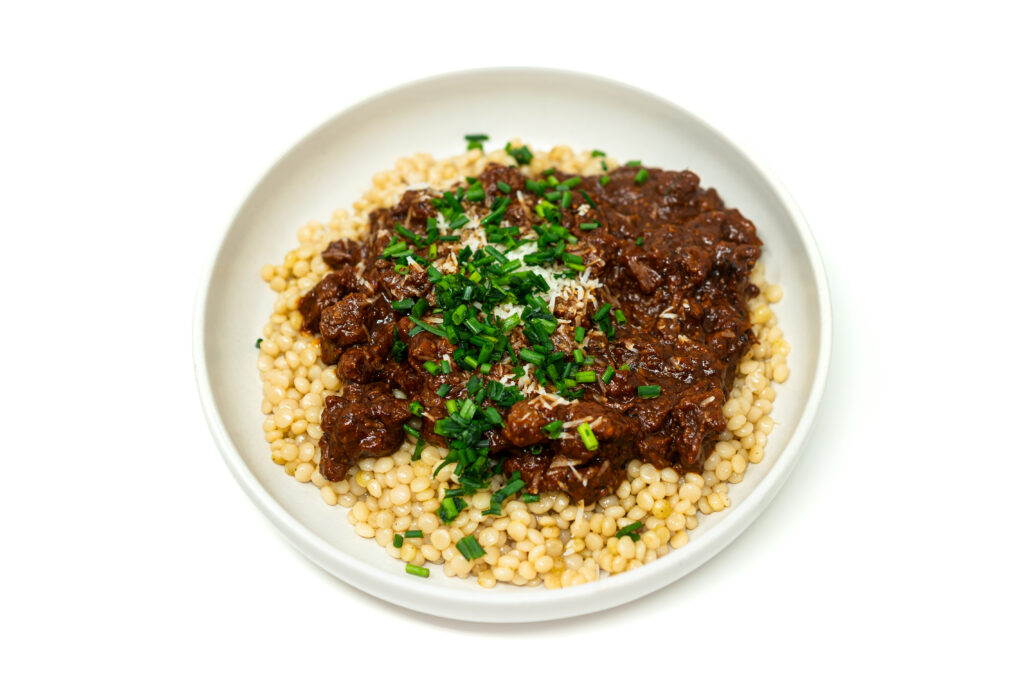 A shallow bowl of tomato soy braised beef brisket served over pearl couscous, topped with chopped chives and Pecorino Romano.