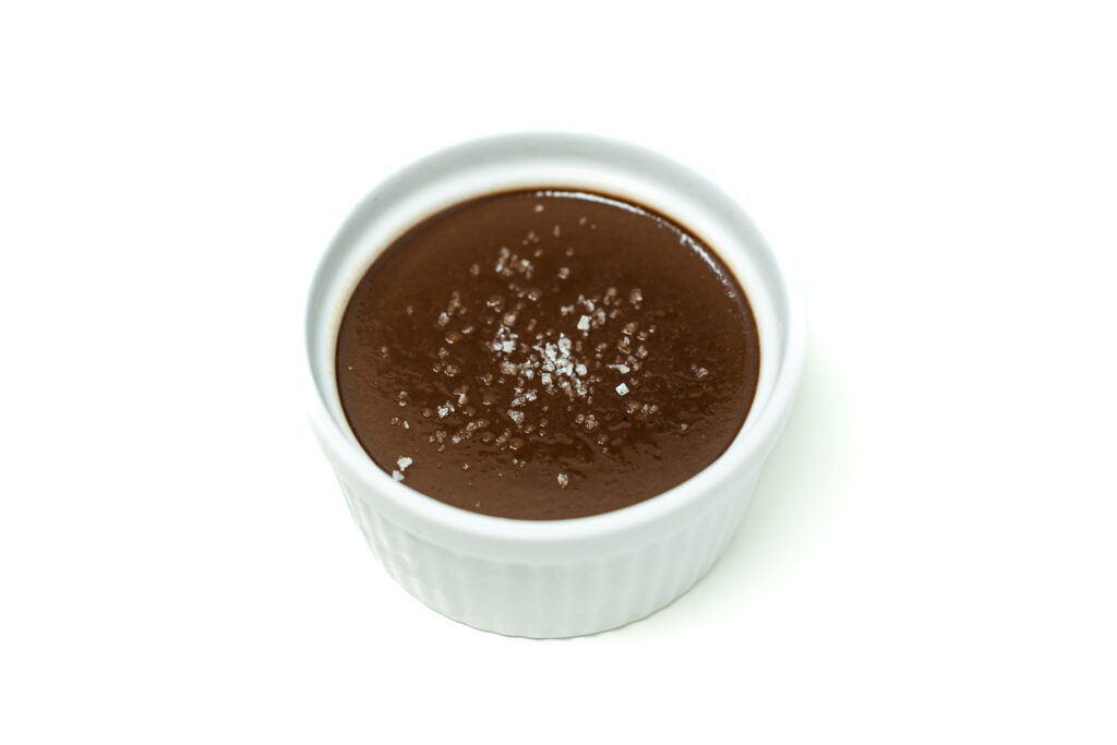 A small ramekin filled with pumpkin cocoa pot de crème, topped with flaky sea salt and a glossy surface reflecting warm light.