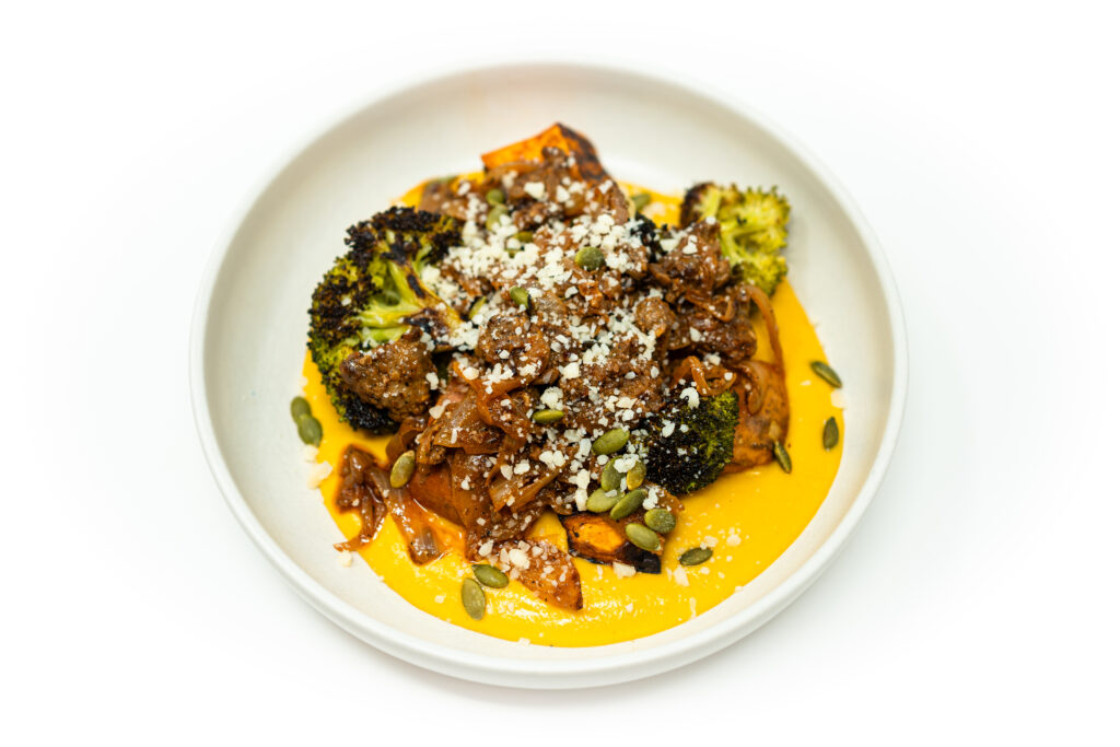 A shallow bowl of French-style ground lamb served over brown butter butternut squash purée with roasted broccoli and sweet potatoes, topped with Parmesan and herbs.