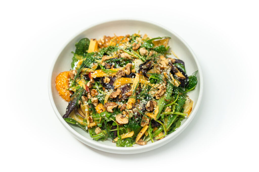 A vibrant salad of caramelized fennel, apples, oranges, mixed greens, and walnuts topped with Parmesan and fennel fronds on a white plate.