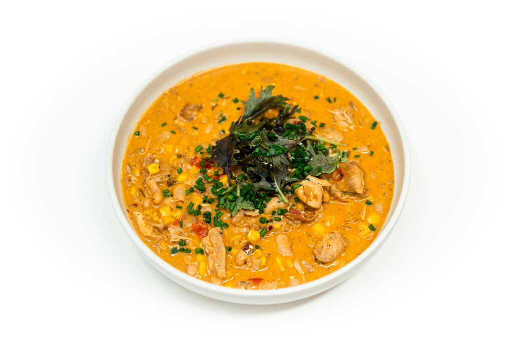 A bowl of creamy buffalo chicken and white bean chili topped with chives and greens on a white background.