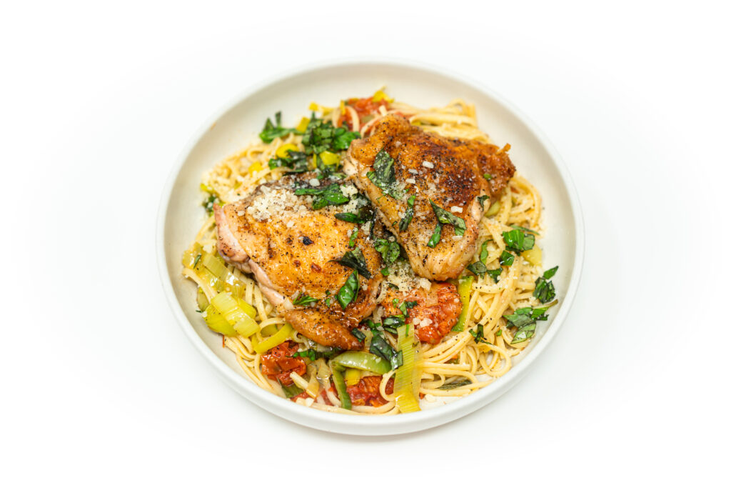 Cast iron chicken thighs with tomato confit pan sauce, leeks, and linguine topped with fresh basil and Parmesan on a rustic plate.