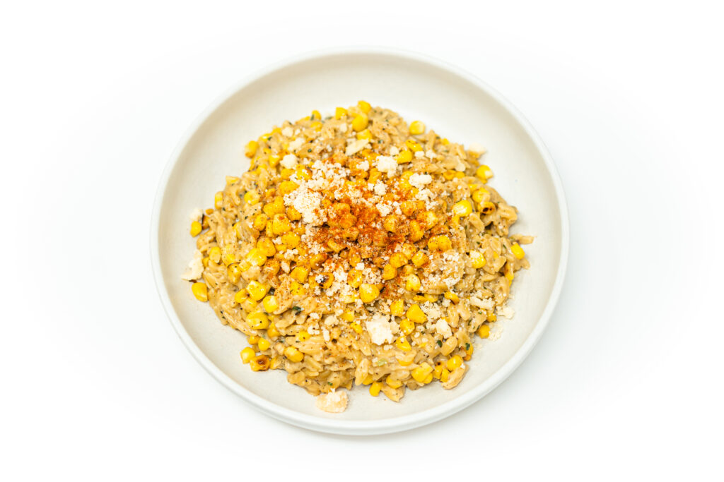 A shallow bowl of creamy corn orzo topped with feta crumbles, charred corn, and fresh chives, glistening under natural light.