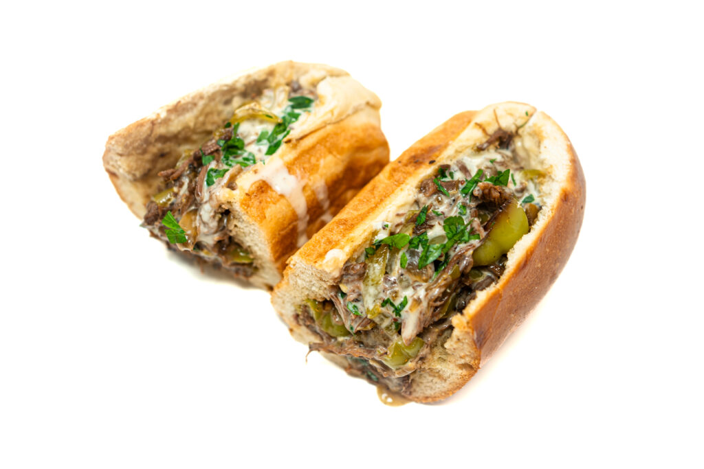 Shredded beef hoagie with blistered peppers and onions, topped with white cheddar cheese sauce on a toasted roll.