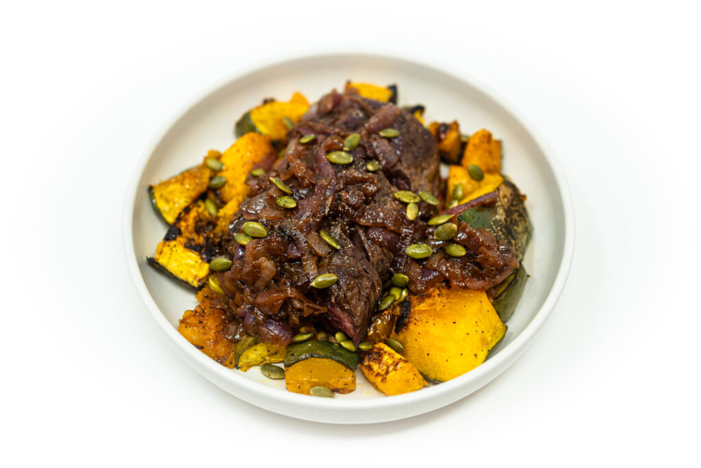 A plate of sliced hanger steak with caramelized onion and rosé shallot sauce served over roasted Jarrahdale pumpkin wedges, topped with toasted pepitas.