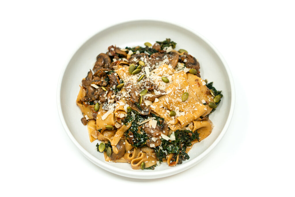 A white bowl filled with pappardelle coated in a mushroom and sun dried tomato sauce with sautéed kale, Parmesan, and toasted pepitas.