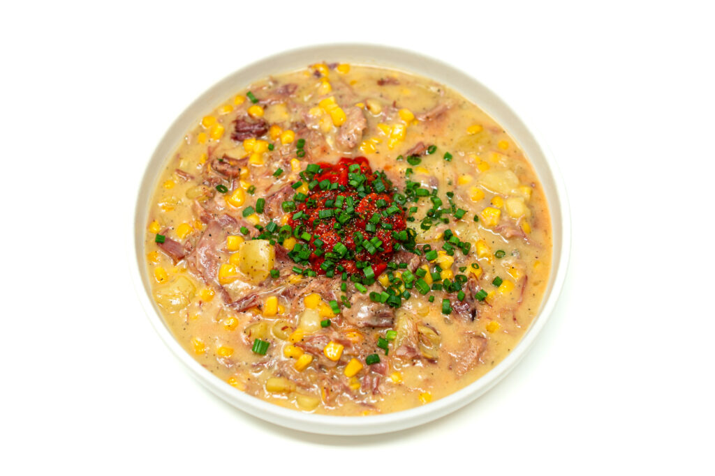 A bowl of smoked ham hock corn chowder topped with fresh red pepper herb salsa surrounded by herbs and ingredients on a rustic table.