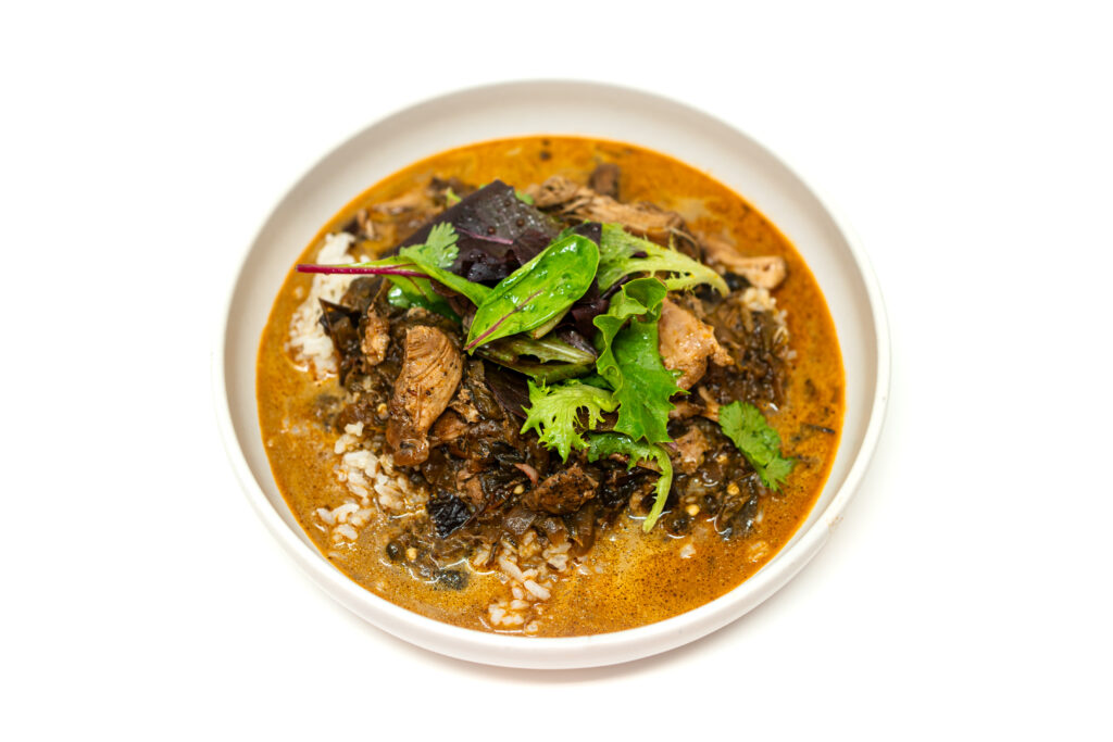 A bowl of smoky poblano and pasilla chicken stew with rice, topped with mixed greens and cilantro in a rich, reddish-brown broth.