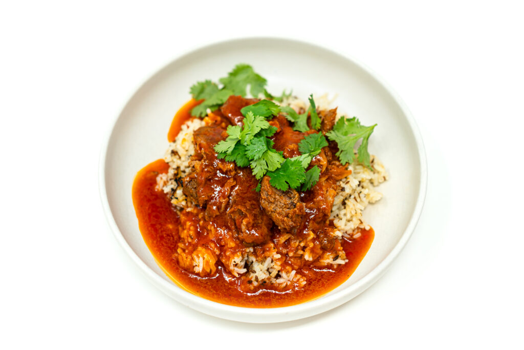 A bowl of tender tomato cumin braised lamb served over toasted cumin jasmine rice, topped with fresh cilantro leaves.
