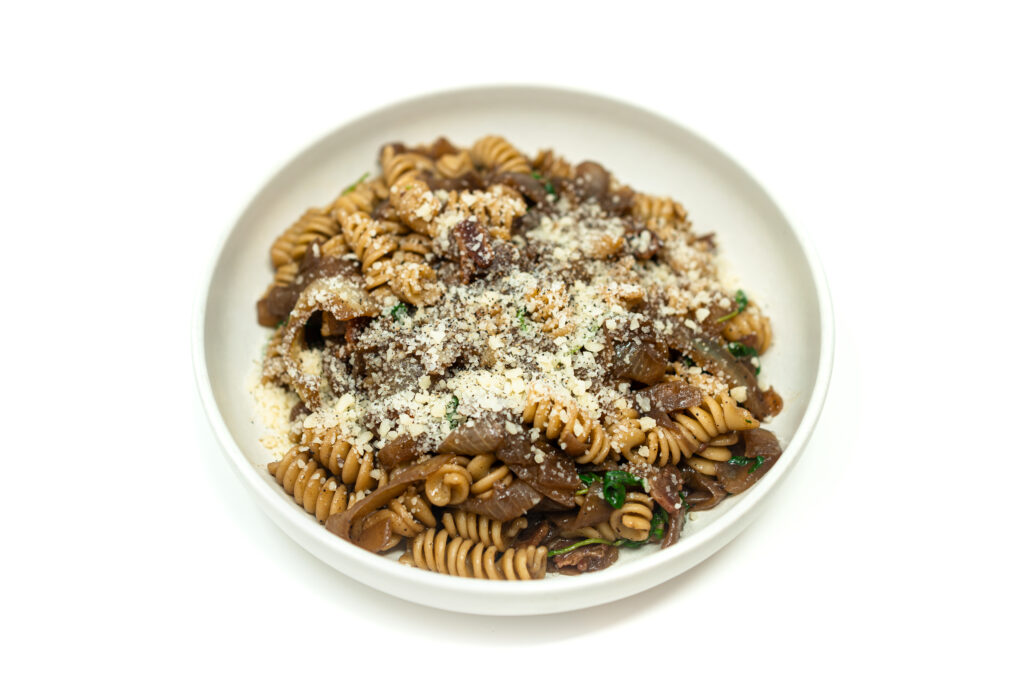 A bowl of red wine pumpkin rotini pasta with bacon, caramelized onions, and arugula, topped with grated Parmesan cheese.