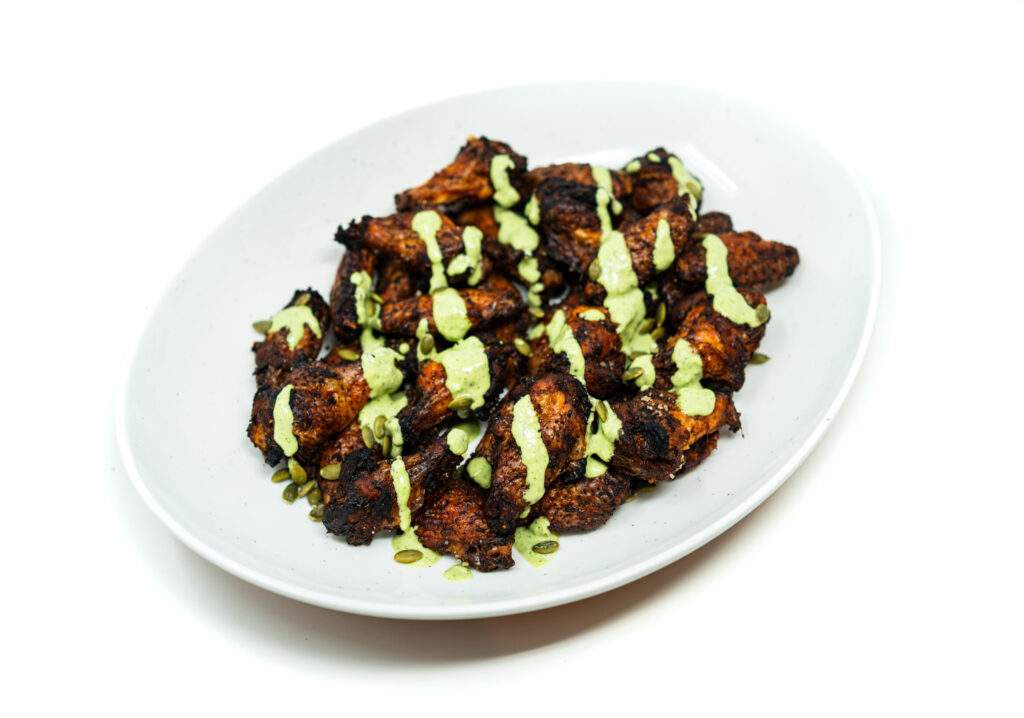 Close-up of jerk-rubbed chicken wings with creamy jalapeño herb sauce, garnished with herbs and lime zest on a rustic plate.