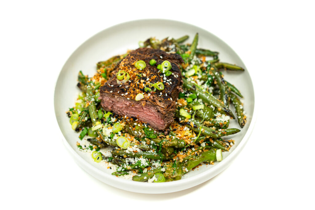 Grilled and sliced New York strip steak layered over a Caesar salad with charred shishito peppers, green beans, arugula, and toasted panko.