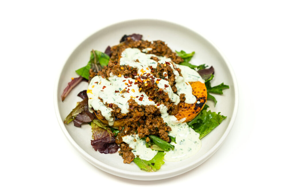 Overhead shot of a white bowl filled with charred butternut squash rounds, ground lamb kefta, and mixed greens, topped with a creamy white herb yogurt sauce and red pepper flakes.